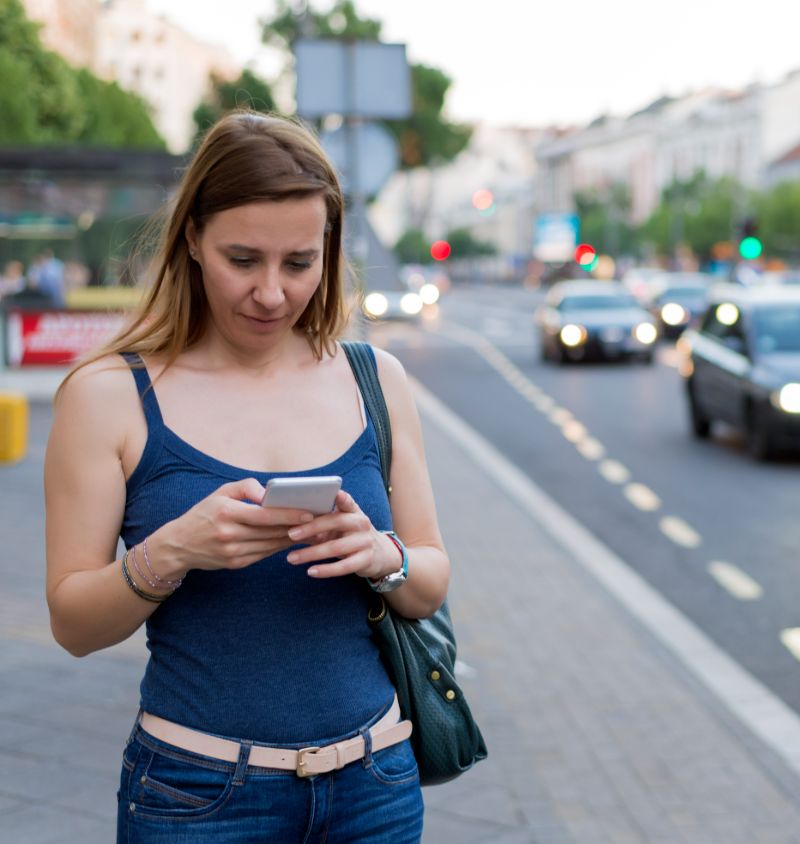 Mulher na calçada usando o celular, com carros passando na avenida ao fundo.
